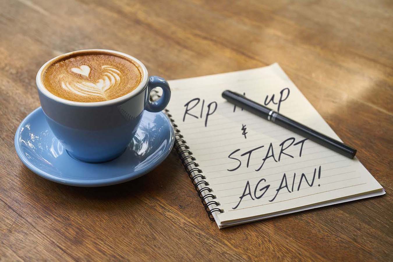wooden table with a cup of cofee beside a notepad with a pen on top, written on pad is Rip it up and start again
