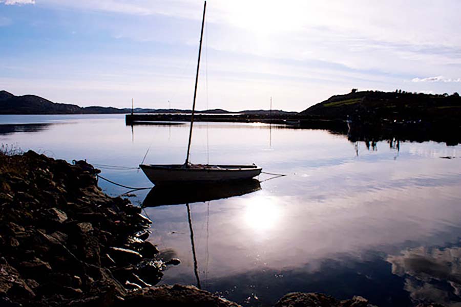 A small boat in silhouette docked on a still water that reflects the sunny sky above