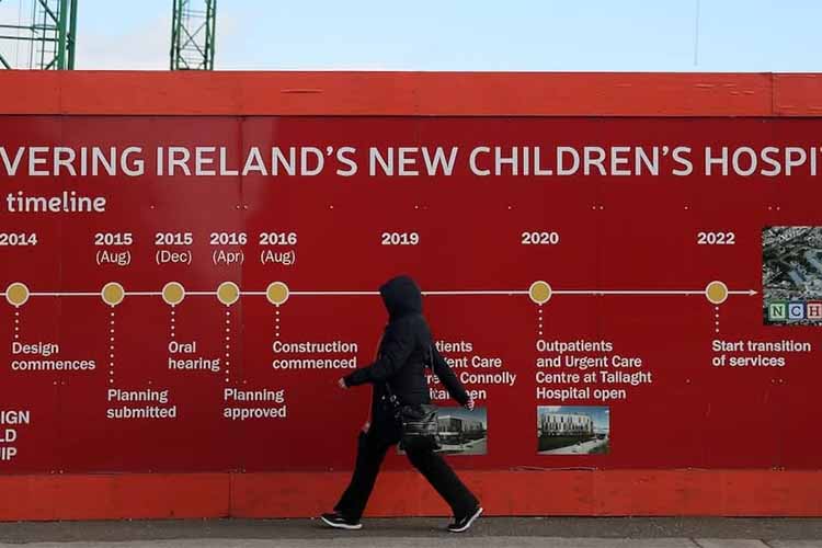 Woman walking past the NCH construction site and board which show its original timeline