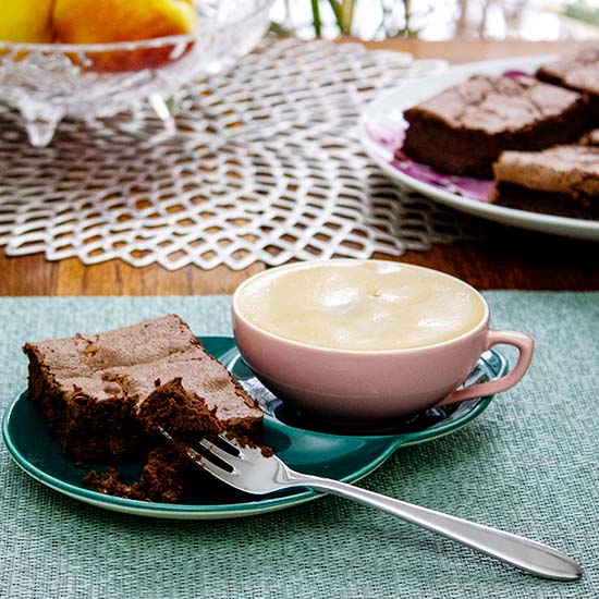 gluten free mocha brownie on a green saucer with a pink cup of foamy coffee, with a plate of mocha brownies in the background with a bowl of apples.